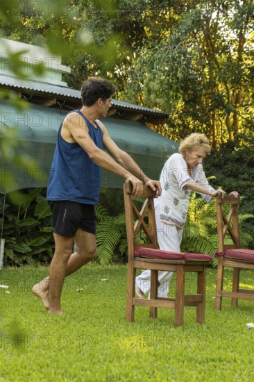 Young male physiotherapist assisting a senior woman performing rehabilitation exercises using chairs in a lush green garden, promoting mobility and recovery