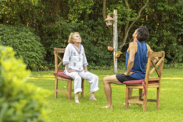 Senior woman and her physiotherapist are sitting on chairs in a garden, doing neck stretching exercises, looking up, as part of a rehabilitation program