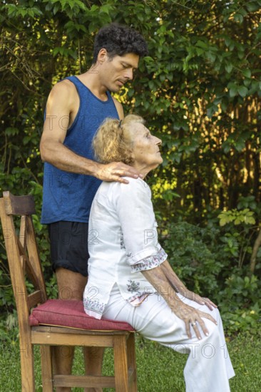 Physiotherapist is guiding a senior woman through posture exercises, providing support and gentle adjustments as she sits on a chair in a serene outdoor setting, promoting mobility and well-being