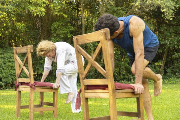 Senior woman and her personal trainer practicing a fitness routine using chairs as support in a backyard setting, promoting healthy aging and mobility