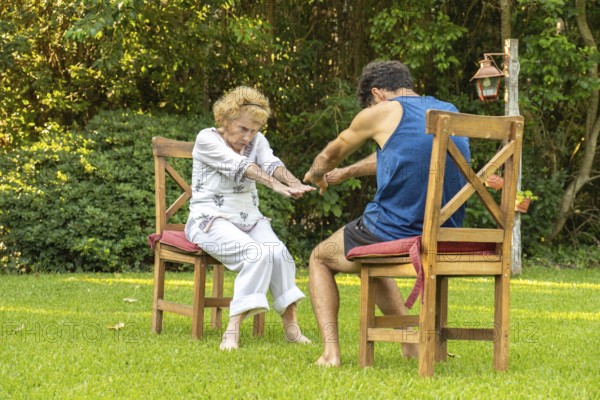 Elderly woman practicing physiotherapy with a personal trainer, performing exercises while sitting on chairs in a garden, promoting health and mobility in old age
