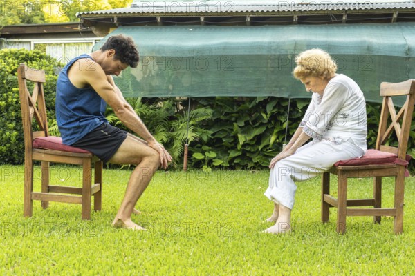 Senior woman and her physiotherapist are performing physical therapy exercises sitting on chairs in a garden, focusing on rehabilitation and improving mobility