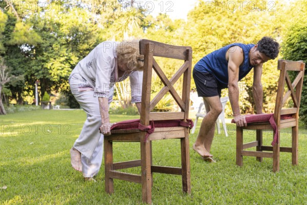 Senior woman and her physiotherapist are performing a rehabilitation exercise using chairs in a garden, promoting mobility and strength in a natural setting