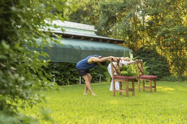 Senior couple practicing stretching exercises in their garden, using chairs as support, promoting flexibility and well-being in a natural environment