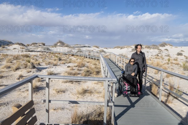 Man and handicapped woman in wheelchair stroll down the Interdune Boardwalk with their ped dog at White Sands Nationl Park near Alamogordo, New Mexico