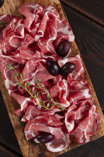 Freshly cut dried meat, served with olives and herbs, on a wooden chopping board, no people
