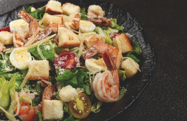 Caesar salad with tiger prawns, shrimp salad, lettuce leaves, quail eggs, cherry tomatoes, parmesan cheese, on a black plate