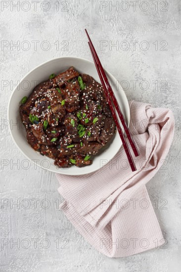 Crispy beef, with green onions and sesame seeds, in a bowl with chopsticks, on a textured surface, on a light background, no people