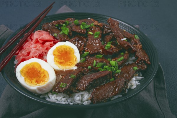 Udon Japanese beef with rice, pickled ginger, boiled egg slice, garnished with green onions, in a black bowl, with chopsticks
