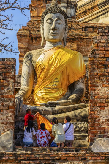 A family decorates a stone Buddha statue with yellow cloth, historic Buddhist temple complex, Ayutthaya, Thailand