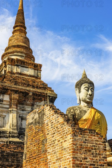 Stone Buddha statue decorated with yellow cloth, in the back a high temple tower, Prang, historic Buddhist temple complex, Ayutthaya, Thailand