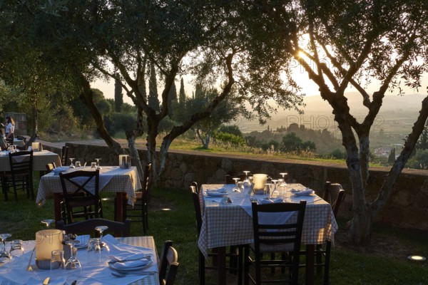 Several outdoor tables set under trees with evening lighting, Hotel Europa, garden restaurant and view of the surrounding landscape, Olympia, Peloponnese, Greece