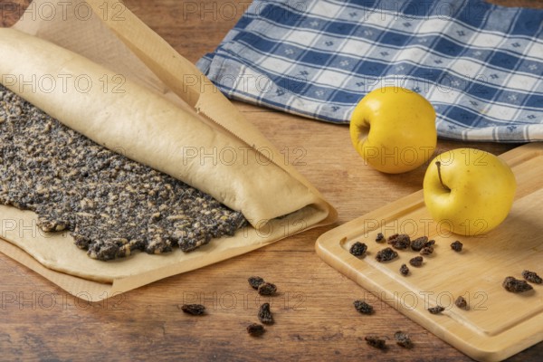 Poppy seed strudel in preparation next to apples and raisins on a wooden table