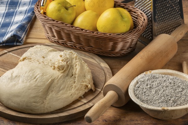Fresh dough lump on board next to bowl with poppy seeds, apples and rolling pin
