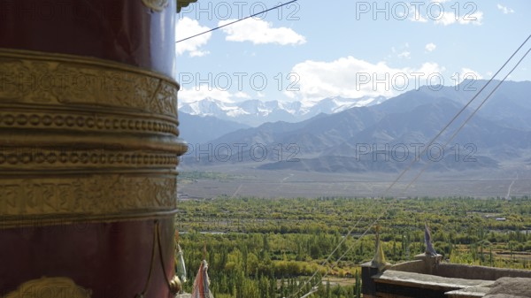 Large prayer wheel in foreground with view of snow-capped mountains and green valley, trekking in Ladakh, Himalayas, India