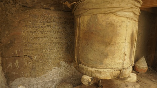 An ancient prayer wheel next to a wall full of inscriptions in a Buddhist context, trekking in Ladakh, Himalayas, India