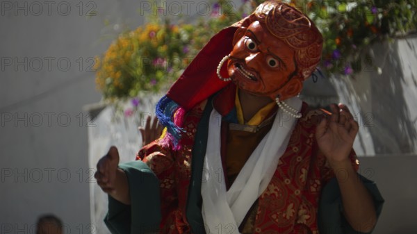 A masked dancer in shades of orange and red with flowers in the background in Leh, trekking in Ladakh, Himalayas, India