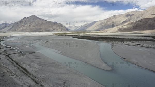 A calm river, Indus flows through a vast landscape surrounded by mountains under a cloudy sky, trekking in Ladakh, Himalayas, India