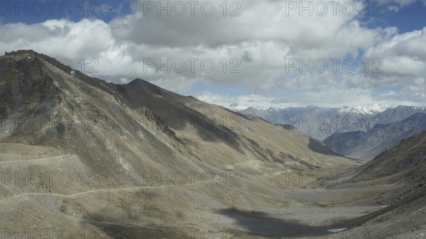 View of a rocky valley with winding mountain road and mountains in the background under slightly cloudy sky, trekking in Ladakh, Himalayas, India