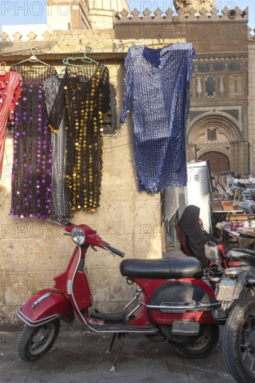 A cool looking red scooter parked amongst a busy street market selling colourful women's dresses near the famous Khan el Khalili Bazaar, historical Cairo, Egypt