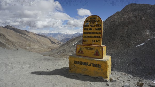 Signpost on dusty Khardung-la Pass in the mountains with sweeping views of surrounding mountains and cloudy skies, trekking in Ladakh, Himalayas, India