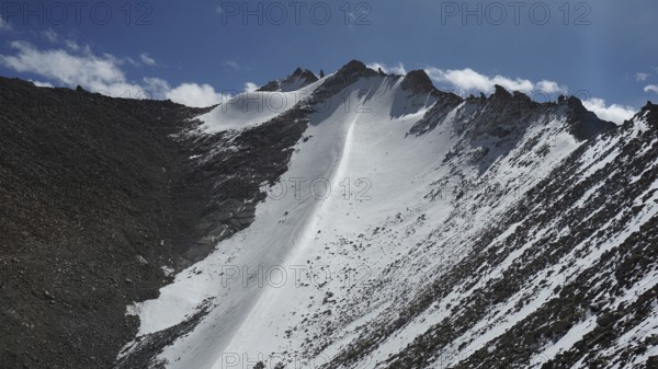 Snowy mountain peak at Khardung-la Pass under clear blue sky with some clouds and rocky landscape, trekking in Ladakh, Himalayas, India