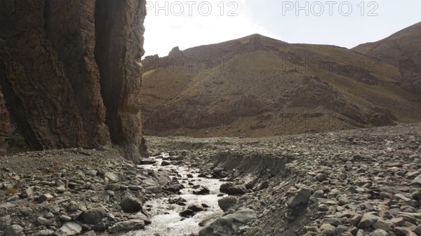 A stream flows through a dry, rocky landscape with high rock walls, trekking at Stok La Pass in Ladakh, Himalayas, India