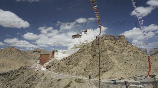 A mountain monastery perches on a rocky hill under a clear sky with floating prayer flags, Ladakh, Himalayas, India