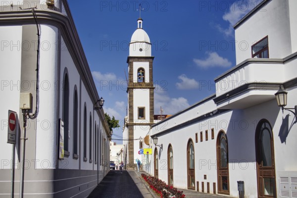White buildings with church tower of the Iglesia de San Ginés church along a paved street under blue sky, Arrecife, Lanzarote, Canary Islands, Spain