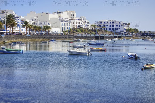 Blue water with boats in the Charco de San Ginés lagoon in front of a coastal town with palm trees and white buildings under clear skies, Arrecife, Lanzarote, Canary Islands, Spain