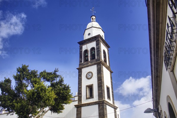 White church tower with clock of the Iglesia de San Ginés church under blue sky, surrounded by trees, Arrecife, Lanzarote, Canary Islands, Spain
