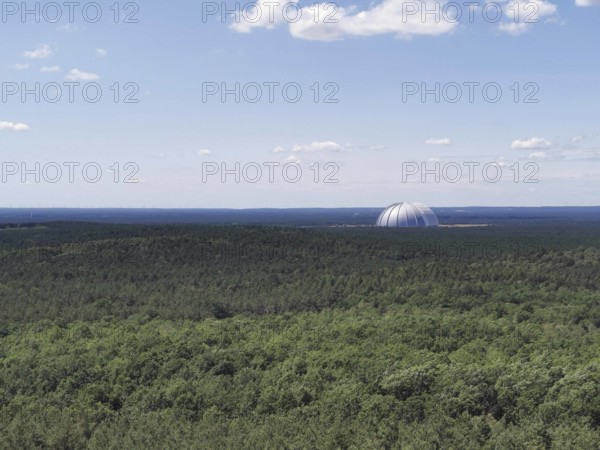 View of an extensive forest with the dome of Tropical Island, Cargolifter Hall on the horizon under a slightly cloudy sky, Spreewald Biosphere Reserve