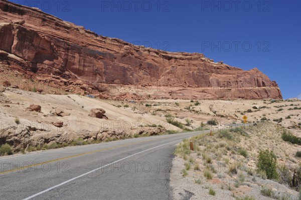 Road leading past a barren, red rock formation under bright blue sky, Arches National Park, Utah, USA
