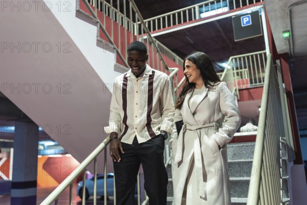 Diverse couple leaving an underground parking lot, laughing and enjoying their date night experience while descending concrete stairs with modern lighting and urban elements