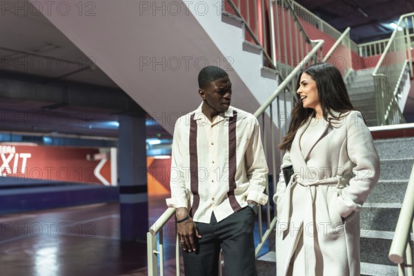 Young diverse couple walking up a concrete staircase in an underground parking facility, engaged in an animated conversation and casual interaction while moving between levels