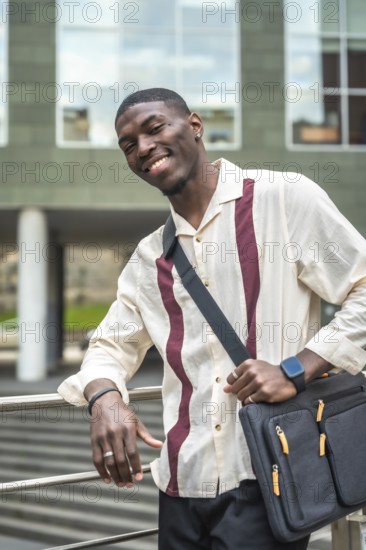 Young black man smiling and posing with laptop bag on his shoulder, leaning against a railing outside a modern building, representing urban lifestyle and professional confidence