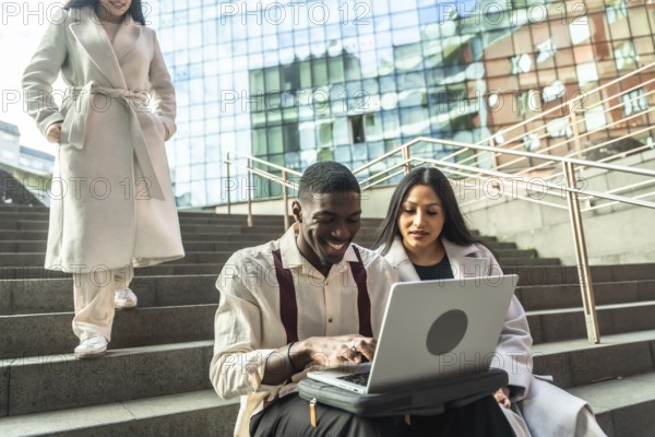 Three diverse business people collaborating outdoors, two using a laptop on city stairs while a third coworker walks past, representing teamwork and modern urban connectivity