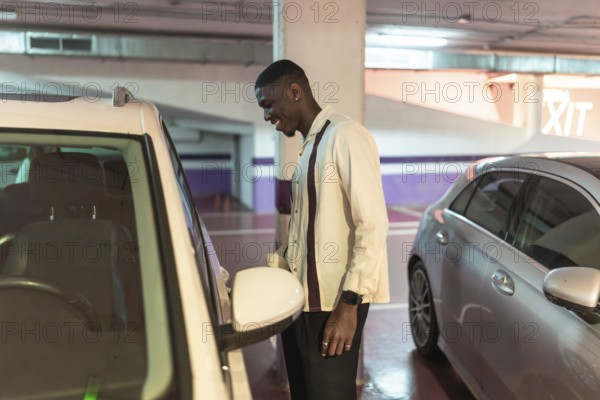 Young black man in an underground parking garage smiling as he opens his white car door, confident and ready for a nighttime urban drive, travel or arrival into the city