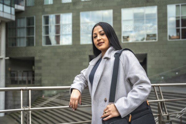Professional latin woman standing by a railing with a messenger bag, looking at the camera against a modern building facade, portraying confidence and urban lifestyle