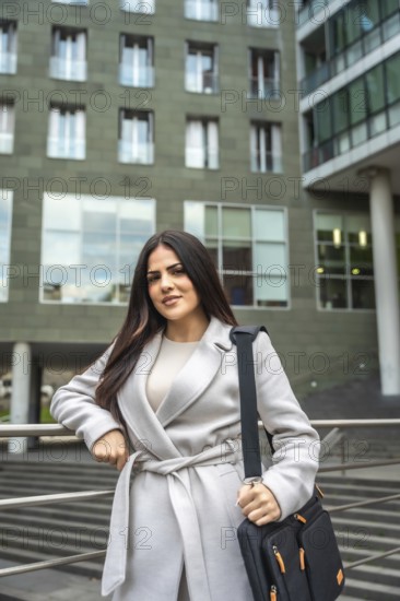 Confident businesswoman wearing a coat and carrying a shoulder bag, standing outdoors in an urban setting with a modern office building behind her, representing success and professionalism