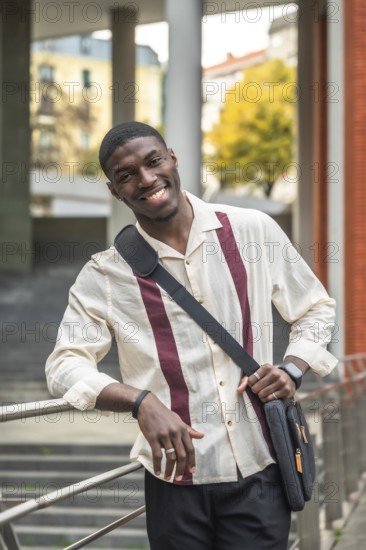 Smiling young black student on a university campus, leaning on a railing with a shoulder bag, confident and optimistic about education, career goals and urban life future