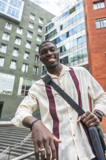 Young black man smiling and leaning on a railing in a modern city street, carrying a bag and dressed business casual, representing confident urban student and young professional life