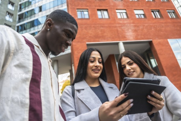 Startup business team including two women and a man collaborating and discussing work using a digital tablet while standing outside modern corporate buildings