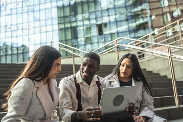 Diverse business professionals or students sitting on urban stairs, discussing project ideas and sharing information on a laptop during an informal outdoor meeting or study session