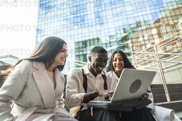 Group of diverse coworkers laughing while sharing content and collaborating on a laptop during an outdoor meeting in an urban setting, reflecting teamwork and success