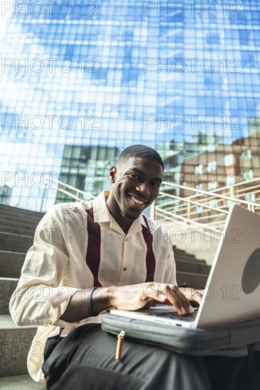 Young black man smiling while typing on a laptop, sitting on urban stairs with a modern glass building reflecting the blue sky in the background, working or studying