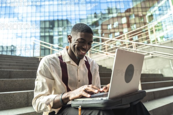 Young black man sitting on concrete stairs in a city, smiling and typing on a laptop, portraying remote work, modern technology, urban lifestyle and professional focus outdoors