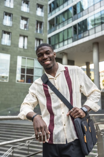 Confident young black professional smiling at camera, leaning on a railing with a messenger bag in a modern city street, smart casual style and upbeat urban vibe