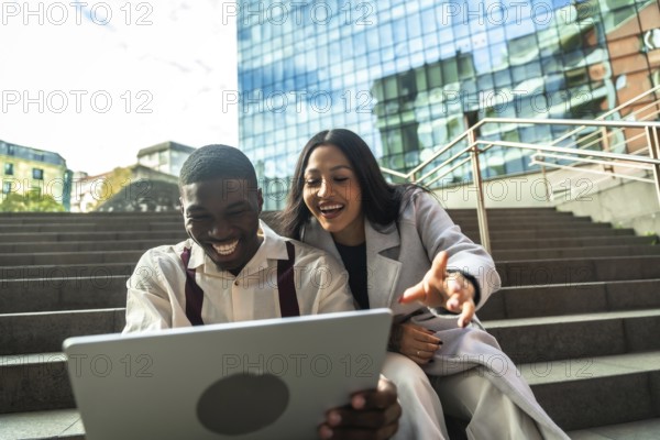 Diverse business colleagues sitting on steps, smiling and laughing while looking at a laptop together, enjoying a break outside a modern office building in the city