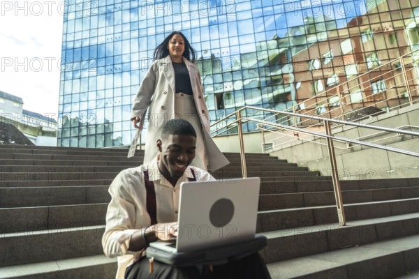 Young professional smiling at laptop on urban steps while a stylish woman walks down nearby by a modern glass building, capturing outdoor remote work and city life ambiance
